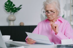 Mulher idosa de óculos examina um documento com expressão de dúvida, sentada à mesa.
