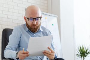 Homem de camisa azul sentado em escritorio lendo atentamente um documento impresso