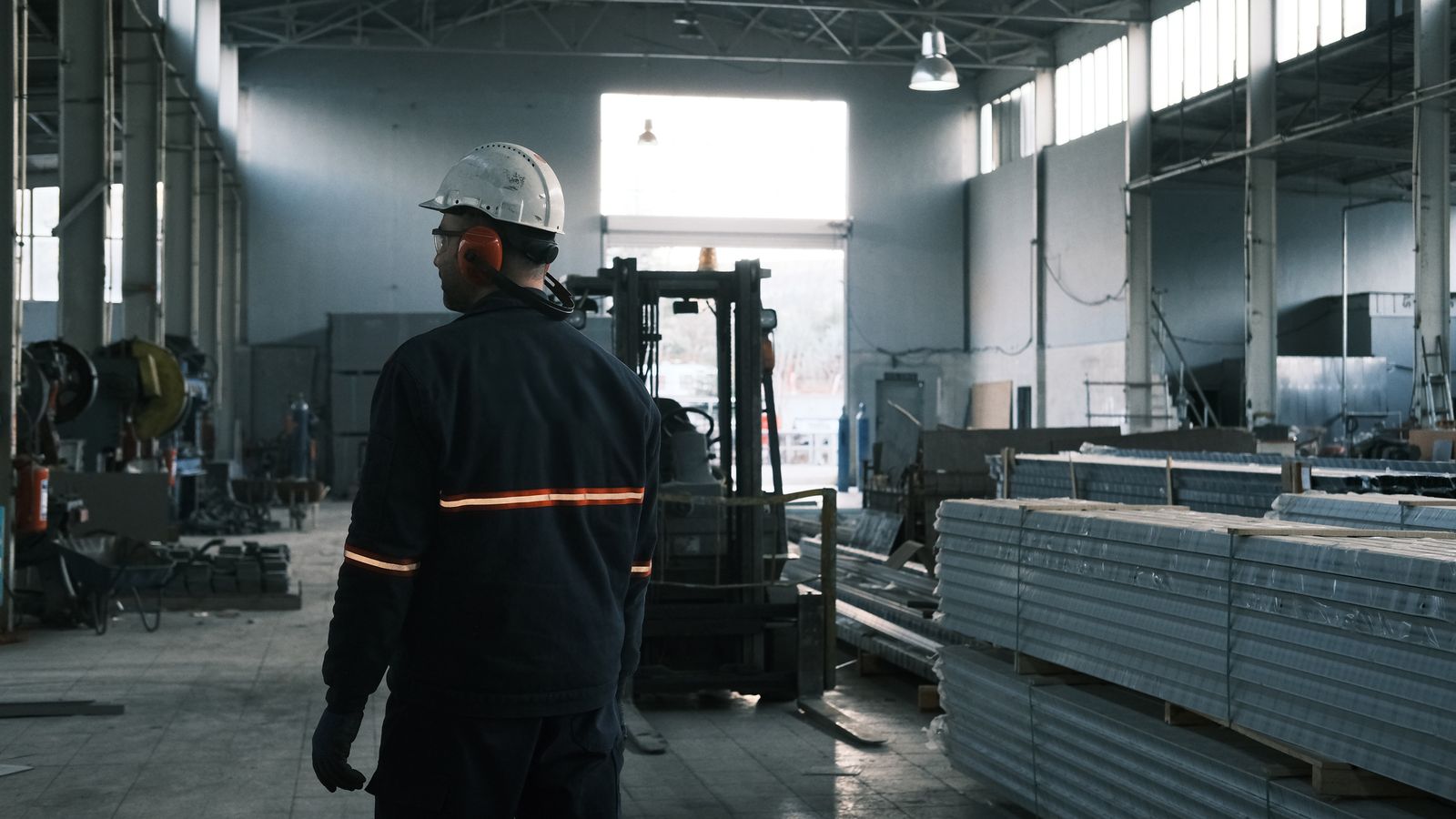 Trabalhador de capacete e protetor auricular de costas em um galpão industrial
