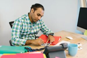 Homem sentado a uma mesa de escritorio conferindo documentos com uma calculadora