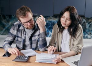 Casal sentado à mesa da cozinha confere contas com calculadora e documentos