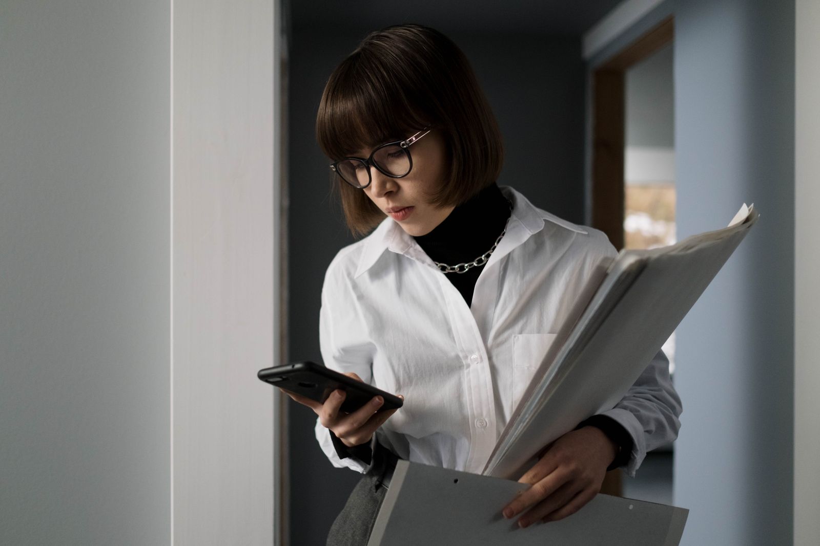 Mulher de óculos segurando um maço de documentos e conferindo uma mensagem no celular