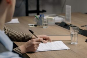 Maos de uma pessoa assinando um documento sobre uma mesa de madeira, com outra pessoa sentada a frente