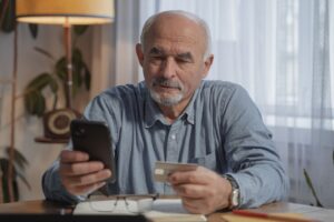 Aposentado confere o celular segurando um cartão à mesa de casa