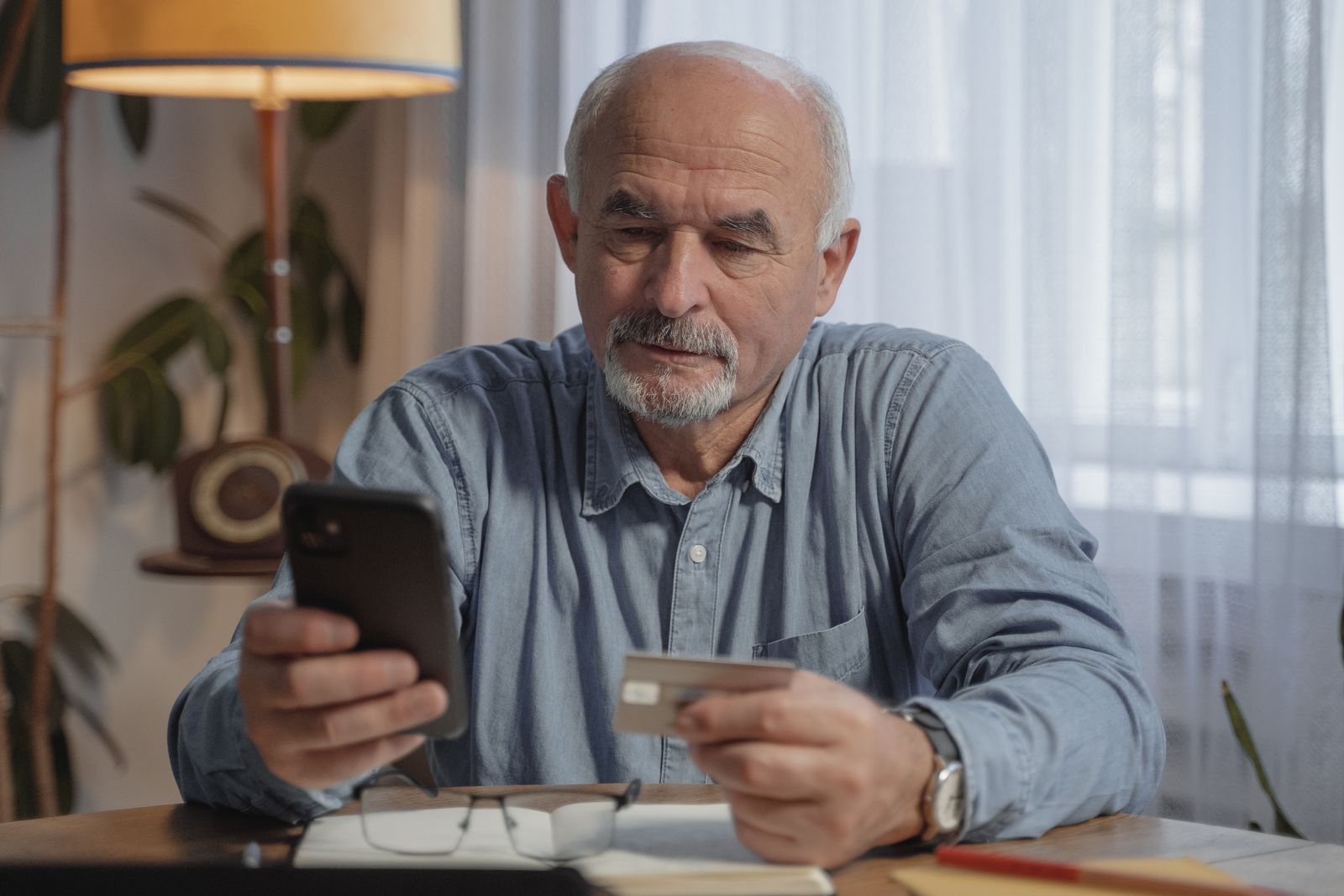 Aposentado confere o celular segurando um cartão à mesa de casa