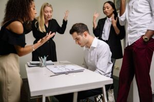 Homem sentado a mesa de reuniao com a cabeca baixa enquanto quatro colegas em pe gesticulam e falam ao mesmo tempo