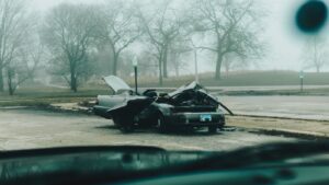 Carro danificado parado à beira de uma rua em dia de neblina, visto de dentro de outro veículo.