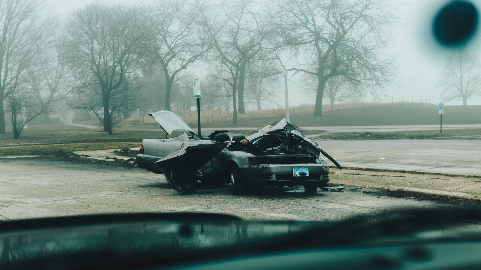 Carro danificado parado à beira de uma rua em dia de neblina, visto de dentro de outro veículo.