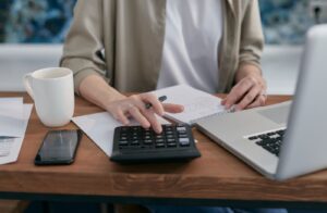 Pessoa usando calculadora ao lado de laptop, bloco de anotações e contas sobre a mesa