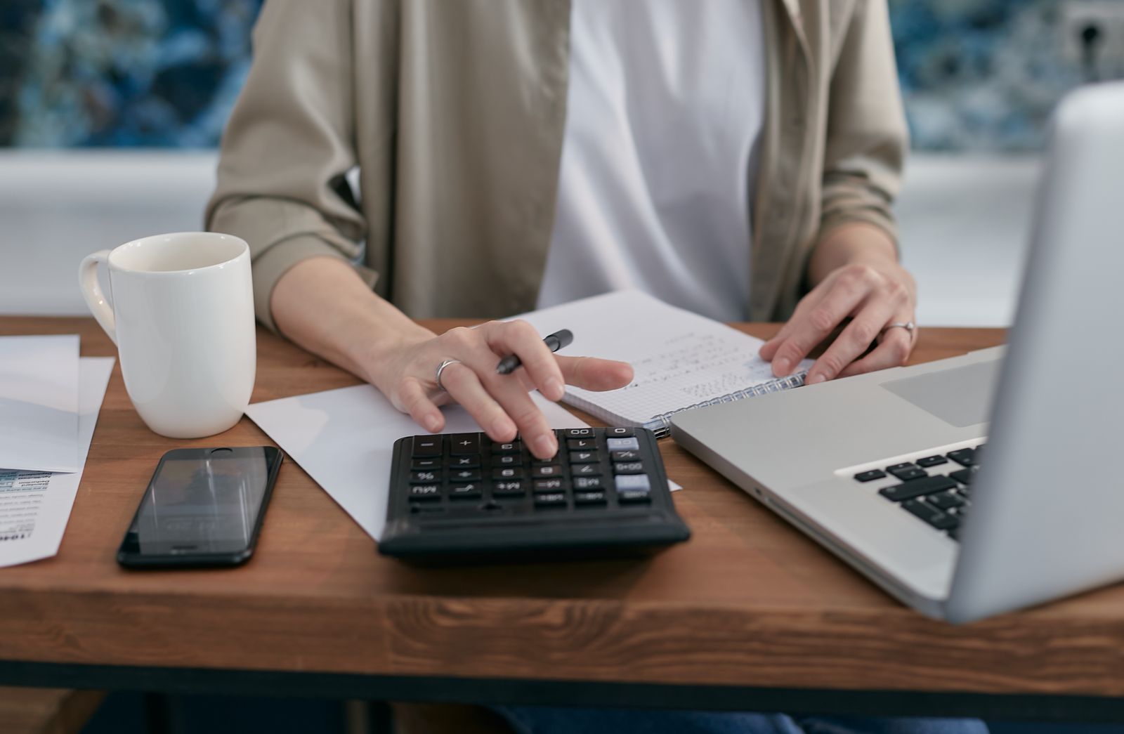 Pessoa usando calculadora ao lado de laptop, bloco de anotações e contas sobre a mesa