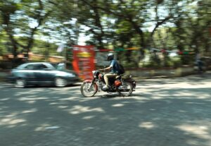 Homem de capacete pilota motocicleta em rua movimentada, com carro passando ao fundo